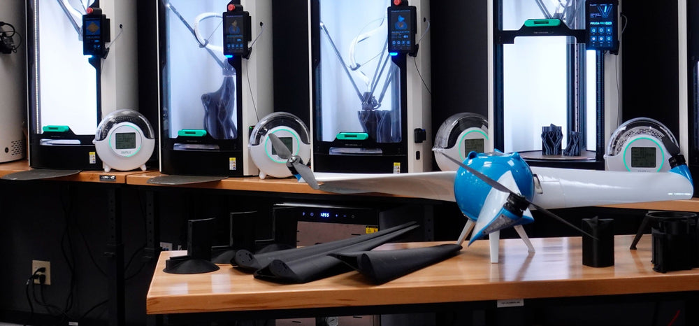 Part of Angel Aerial's five-printer Prusa Pro HT90 fleet running beside the assembled Trio airframe
