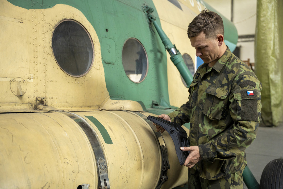 A soldier in camouflage uniform with a Czech Republic flag patch holds a large black polymer part with a bolted circular hub, beside the riveted yellow and green fuselage of a military helicopter in a hangar.