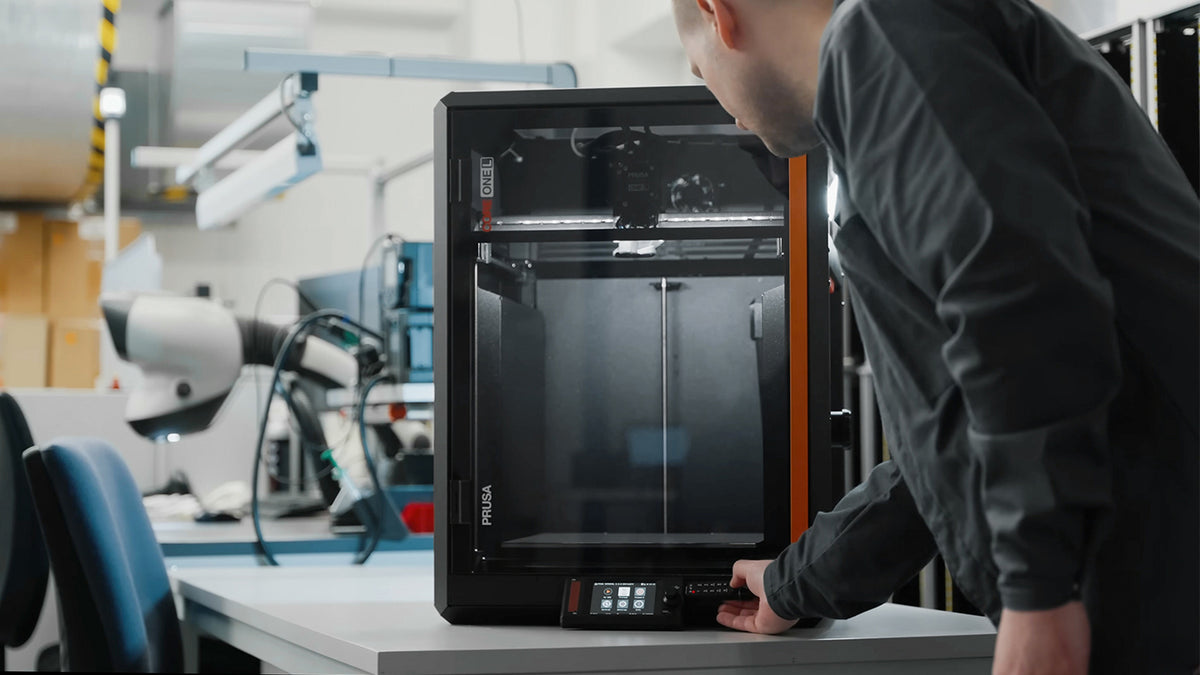 A person at a workbench rests one hand on the front control panel of a black enclosed Prusa CORE One L 3D printer; benches, shelving and lab equipment fill the workshop behind.