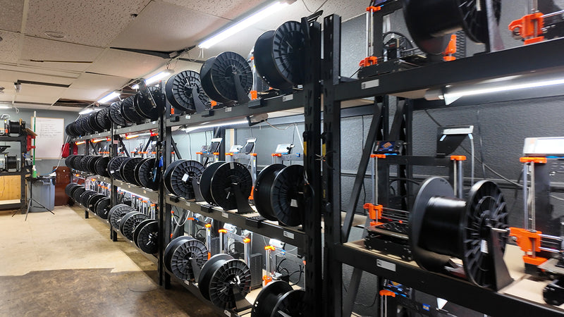 Two long racks of open-frame Original Prusa 3D printers filling a basement print farm, each machine with a black filament spool mounted on the rack beside it, running down a narrow aisle under strip lighting